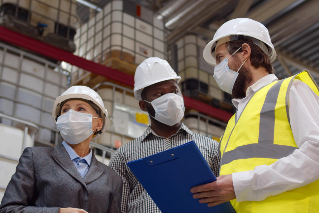 a trio of logistics professionals wearing safety masks while discussing warehouse health and safety measure on clipboard